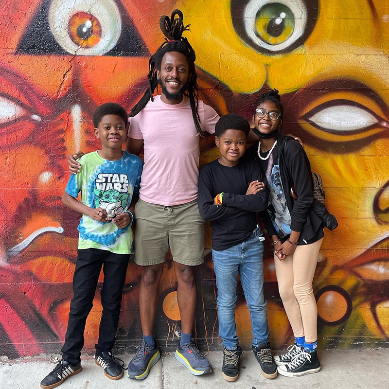 Photo of Mike Simmons and three smiling kids in front of a colorful mural